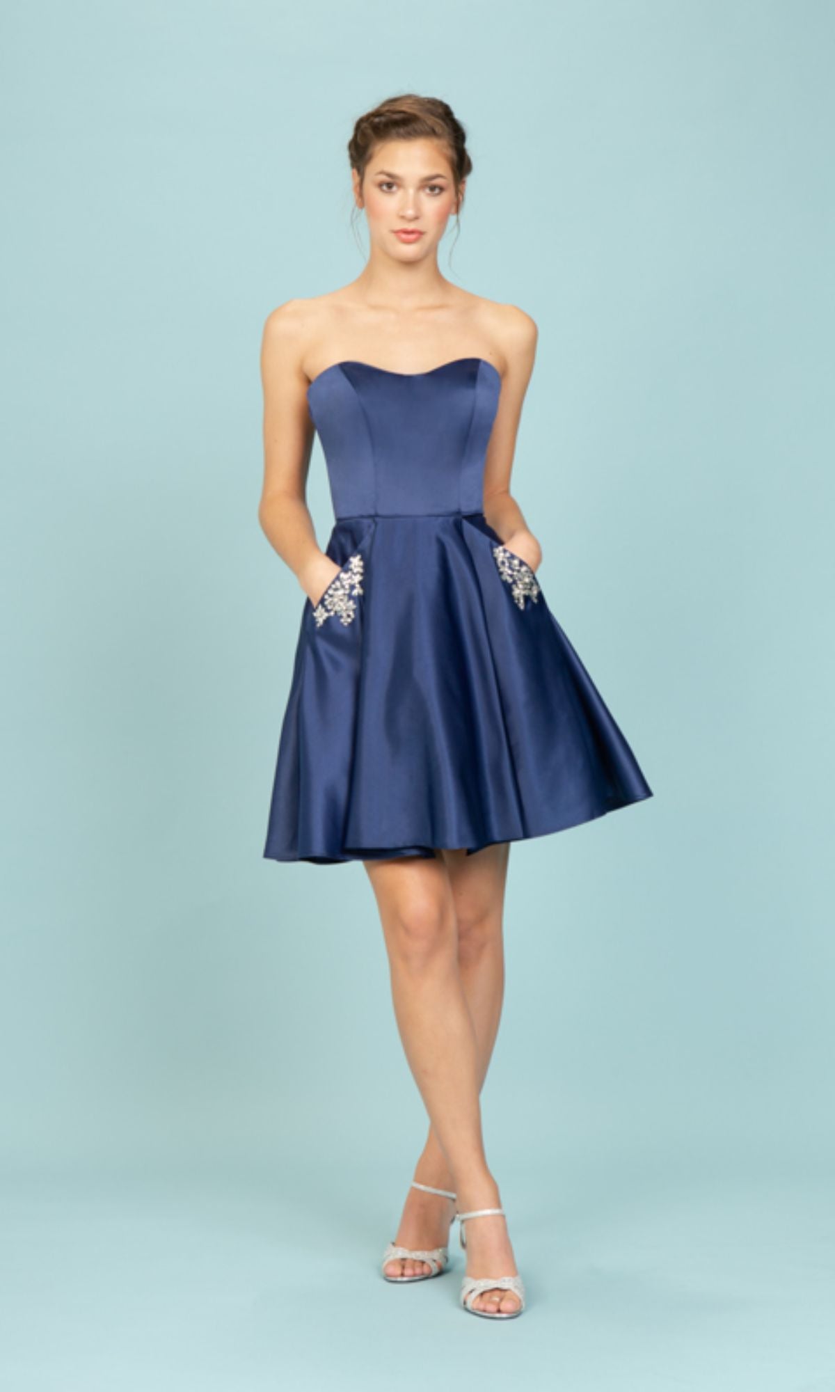A person in the Eureka 6622 Strapless Short Wedding-Guest Dress—knee-length, dark blue with floral embroidered pockets—and silver open-toe heels stands against a light blue background.